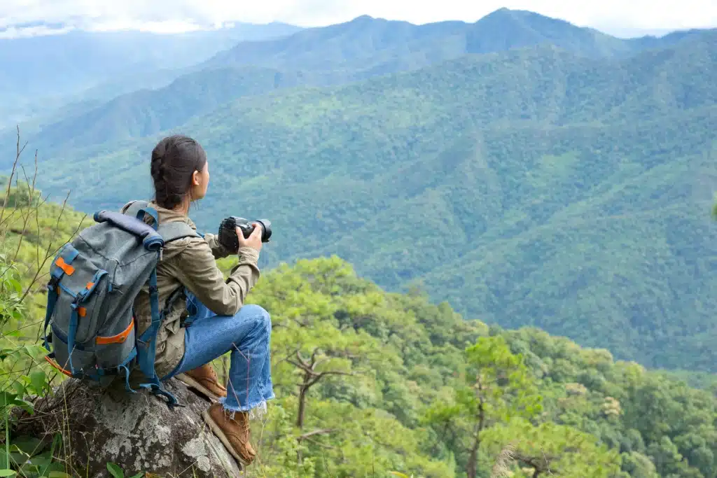A man sitting with a camera facing towards the hills covered with greenery