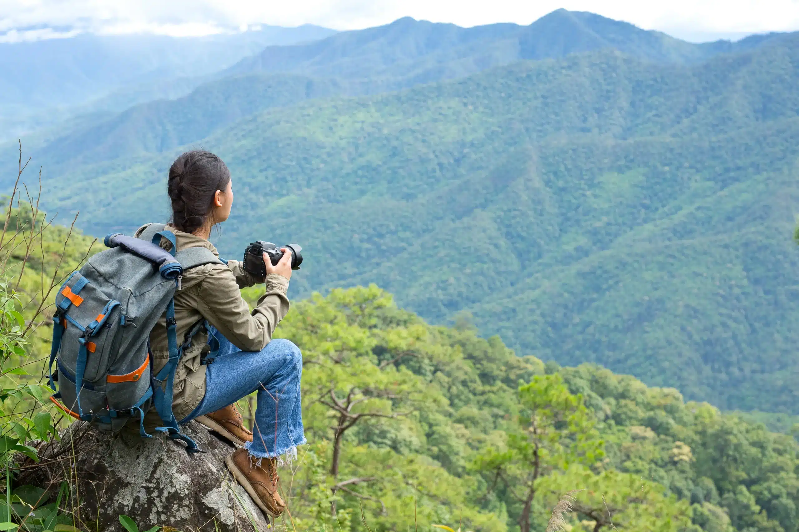 A man sitting with a camera facing towards the hills covered with greenery