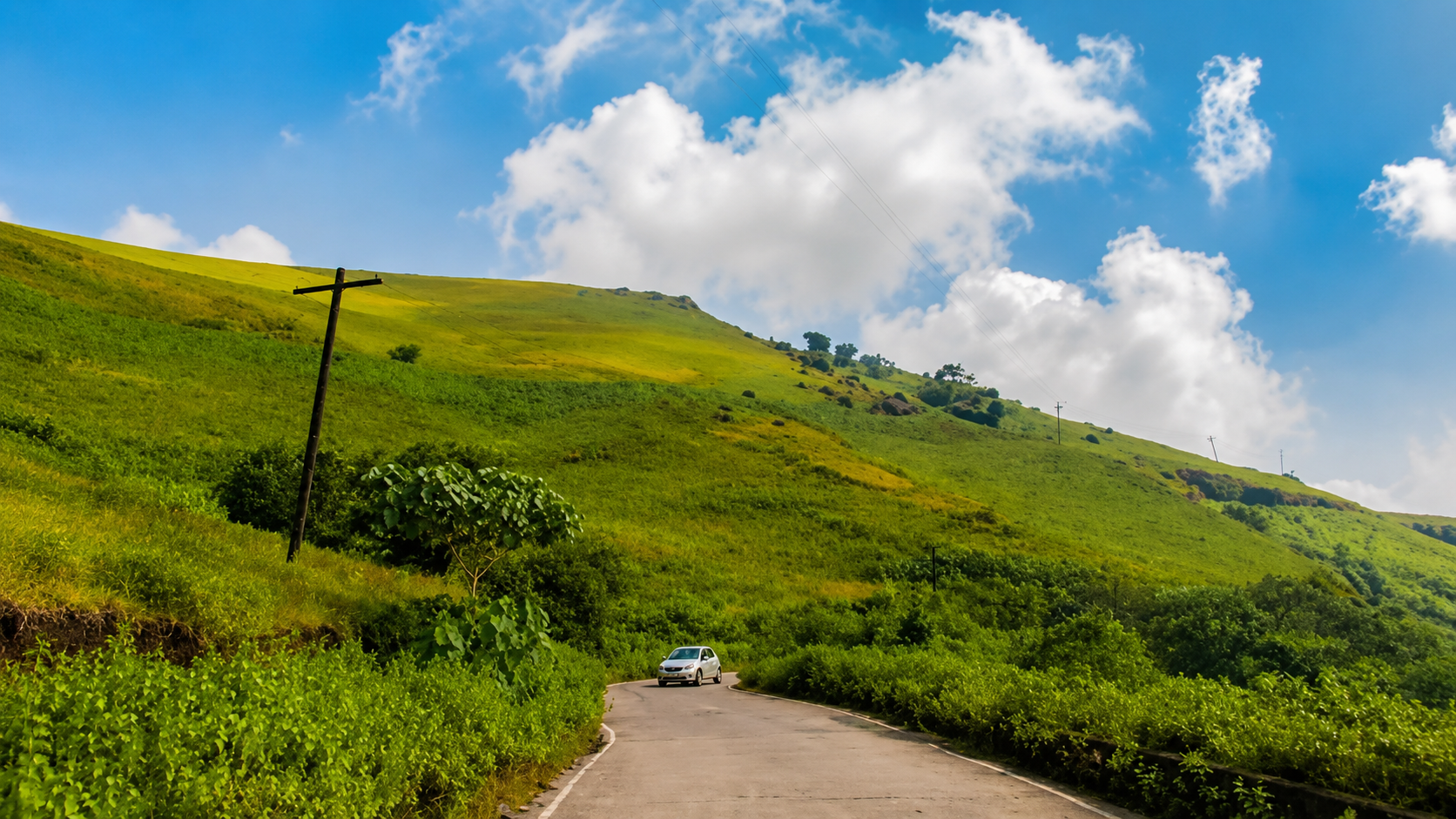 Scenic winding road through lush green hills in Coorg under a bright blue sky with white clouds, creating a peaceful vacation landscape.